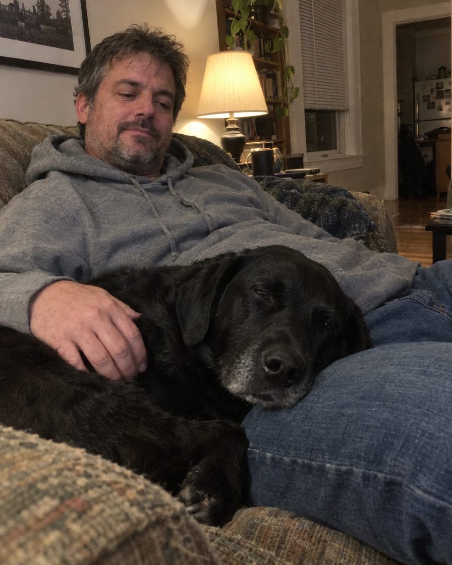 A man relaxing on the couch with his black labrador mix resting its head on his lap